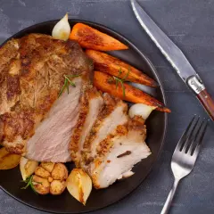 Pieczona szynka z marchewką, cebulą i czoskiem na talerzu, obok nóż i widelec.