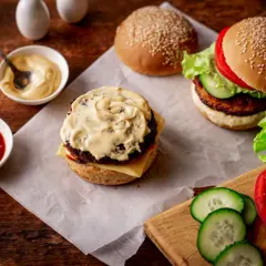 Dwa burgery w trakcie przygotowania, jeden z sosem do burgera, obok miseczki z ketchupem i majonezem, świeże warzywa.