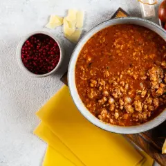 Sos bolognese w misce, obok płaty makaronu, parmezan, pomidorki i pieprz. Idealne składniki, by dowiedzieć się, jak zawekowac sos bolognese.