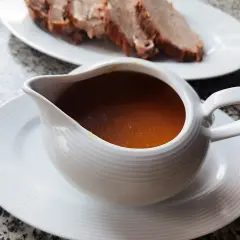 Aromaticzny sos do pieczeni w sosjerce, obok talerz z pokrojonym mięsem. Idealny dodatek, by dowiedzieć się, jak zrobić sos do pieczeni.
