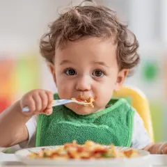 Uroczy maluch w zielonym śliniaczku je posiłek. Pytanie, ile gotować mięso dla niemowlaka, jest kluczowe dla jego zdrowia.