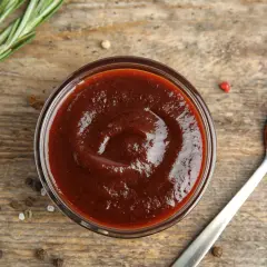 Domowy sos BBQ z wędzonym aromatem bez grilla. Gęsty, ciemny sos w miseczce i na łyżce, obok gałązka rozmarynu i ziarna pieprzu.