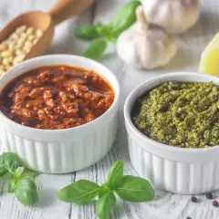 Dwa rodzaje pesto, zielone i czerwone, w białych miseczkach. Obok składniki: orzeszki piniowe, czosnek, bazylia, parmezan. Zastanawiasz się, ile czasu można przechowywać domowe pesto w lodówce?