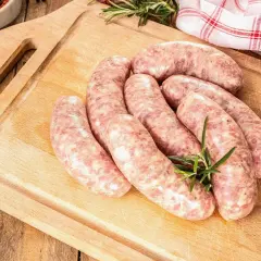 Surowe kiełbaski na desce, gotowe do rozmrożenia. Obok gałązka rozmarynu, czosnek i miseczka z przyprawami.