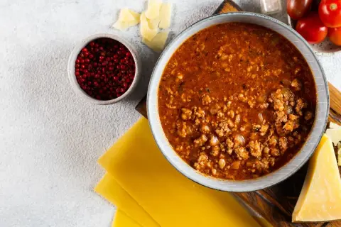 Sos bolognese w misce, obok płaty makaronu, parmezan, pomidorki i pieprz. Idealne składniki, by dowiedzieć się, jak zawekowac sos bolognese.