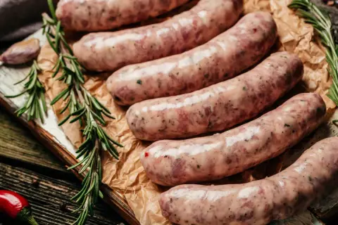 Surowa biała kiełbasa na desce, obok gałązki rozmarynu i papryczki chili. Czy można mrozić białą kiełbasę? Tak, można ją mrozić.