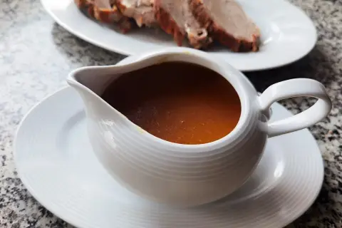 Aromaticzny sos do pieczeni w sosjerce, obok talerz z pokrojonym mięsem. Idealny dodatek, by dowiedzieć się, jak zrobić sos do pieczeni.