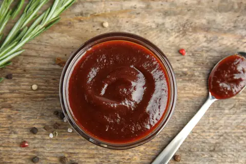 Domowy sos BBQ z wędzonym aromatem bez grilla. Gęsty, ciemny sos w miseczce i na łyżce, obok gałązka rozmarynu i ziarna pieprzu.