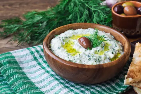Oryginalny przepis na sos tzatziki: kremowy jogurt, świeży ogórek, czosnek, koperek i oliwa, ozdobiony oliwką.
