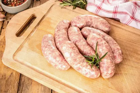 Surowe kiełbaski na desce, gotowe do rozmrożenia. Obok gałązka rozmarynu, czosnek i miseczka z przyprawami.