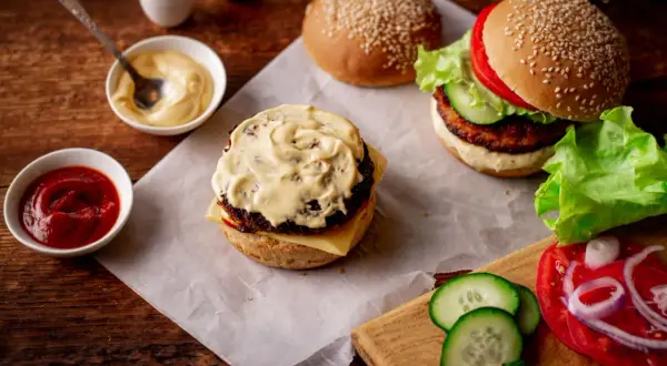 Dwa burgery w trakcie przygotowania, jeden z sosem do burgera, obok miseczki z ketchupem i majonezem, świeże warzywa.