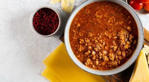 Sos bolognese w misce, obok płaty makaronu, parmezan, pomidorki i pieprz. Idealne składniki, by dowiedzieć się, jak zawekowac sos bolognese.