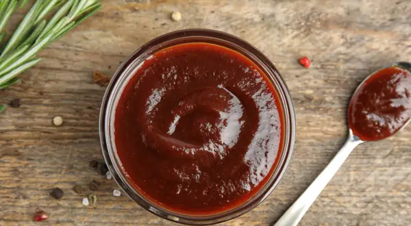 Domowy sos BBQ z wędzonym aromatem bez grilla. Gęsty, ciemny sos w miseczce i na łyżce, obok gałązka rozmarynu i ziarna pieprzu.