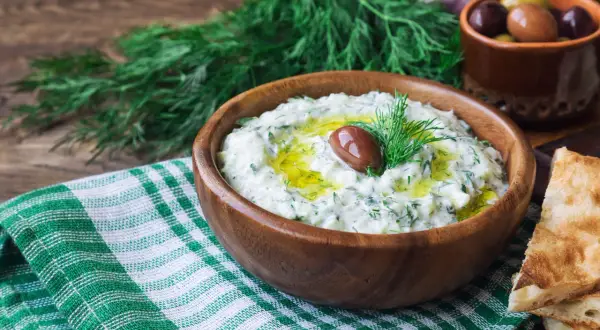 Oryginalny przepis na sos tzatziki: kremowy jogurt, świeży ogórek, czosnek, koperek i oliwa, ozdobiony oliwką.