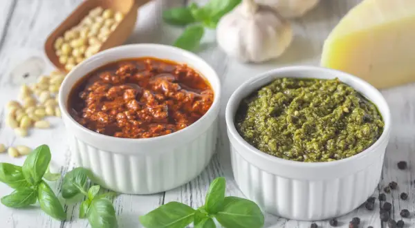 Dwa rodzaje pesto, zielone i czerwone, w białych miseczkach. Obok składniki: orzeszki piniowe, czosnek, bazylia, parmezan. Zastanawiasz się, ile czasu można przechowywać domowe pesto w lodówce?