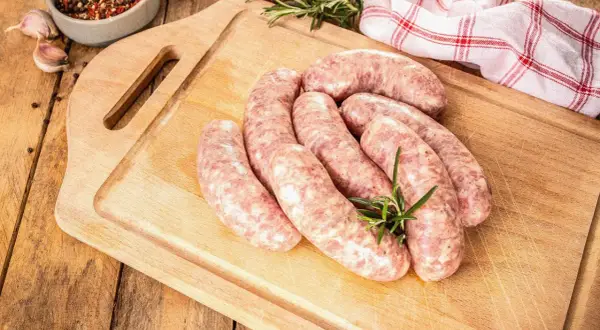 Surowe kiełbaski na desce, gotowe do rozmrożenia. Obok gałązka rozmarynu, czosnek i miseczka z przyprawami.
