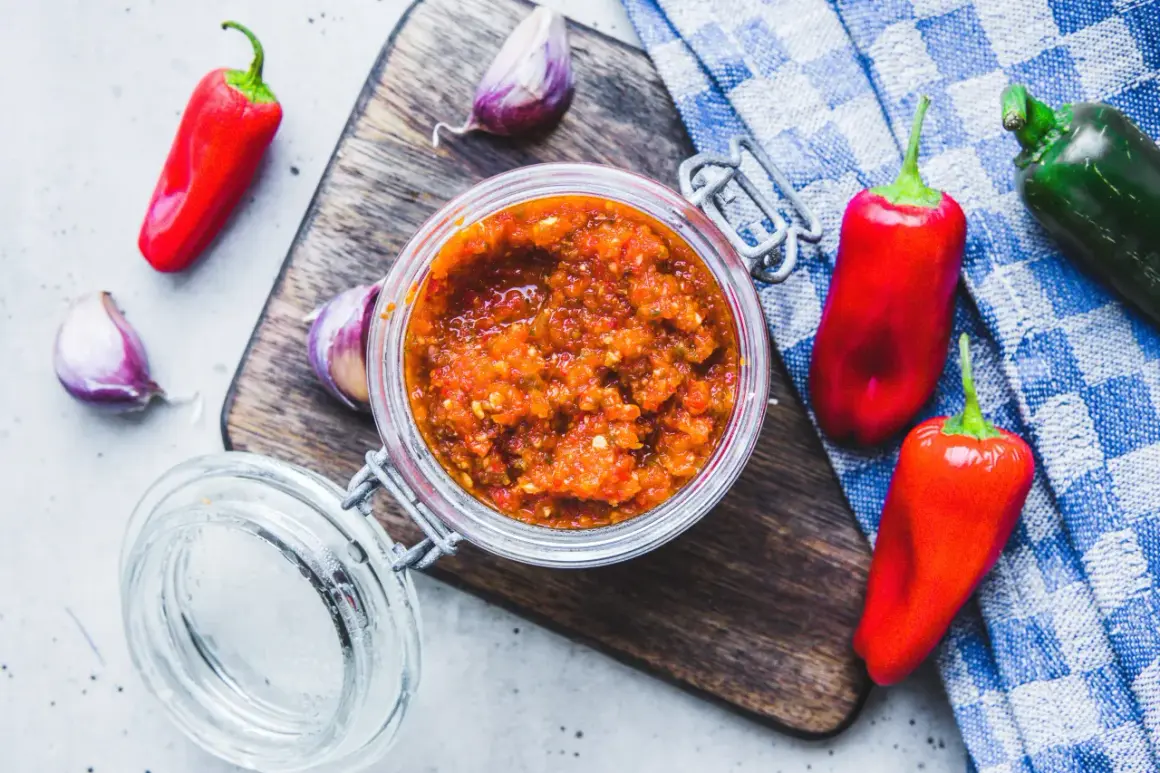 Domowy sos sriracha z papryczek chilli i czosnku w słoiku, otoczony świeżymi papryczkami i czosnkiem na desce.
