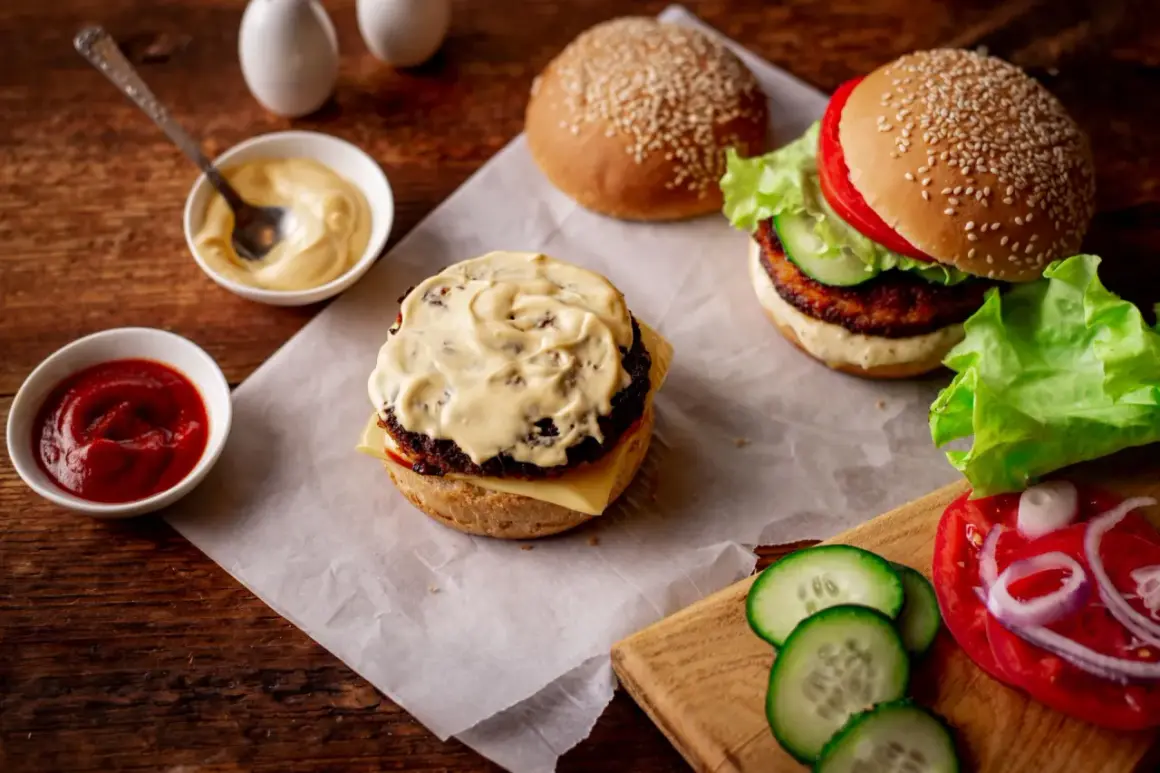 Dwa burgery w trakcie przygotowania, jeden z sosem do burgera, obok miseczki z ketchupem i majonezem, świeże warzywa.
