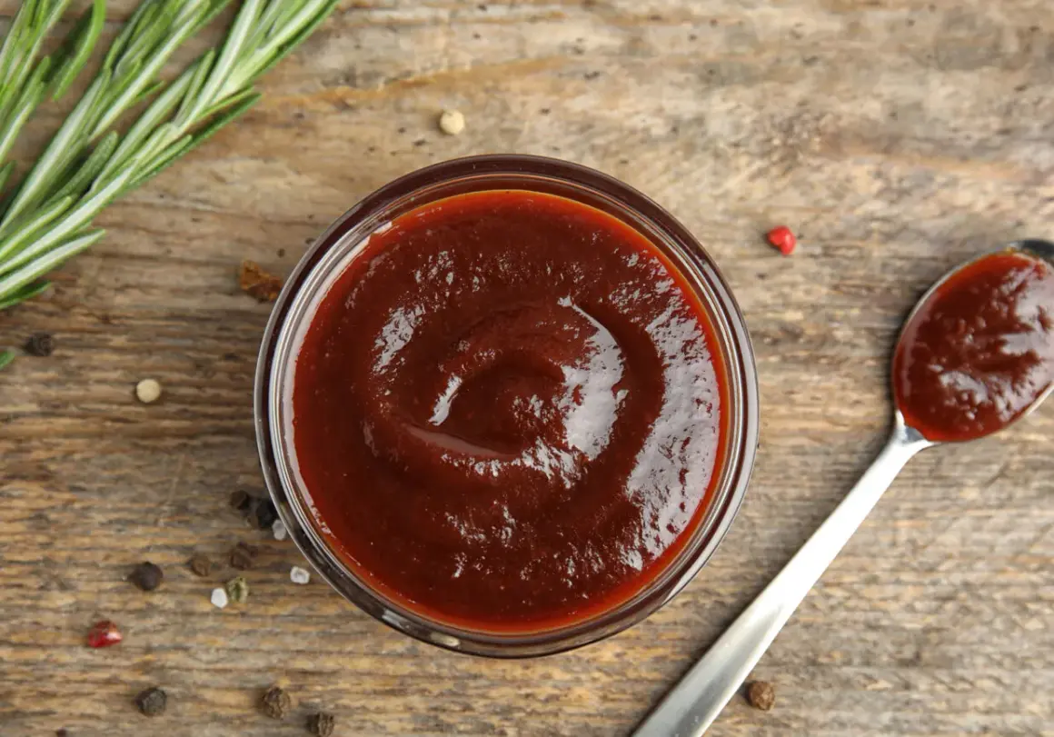 Domowy sos BBQ z wędzonym aromatem bez grilla. Gęsty, ciemny sos w miseczce i na łyżce, obok gałązka rozmarynu i ziarna pieprzu.