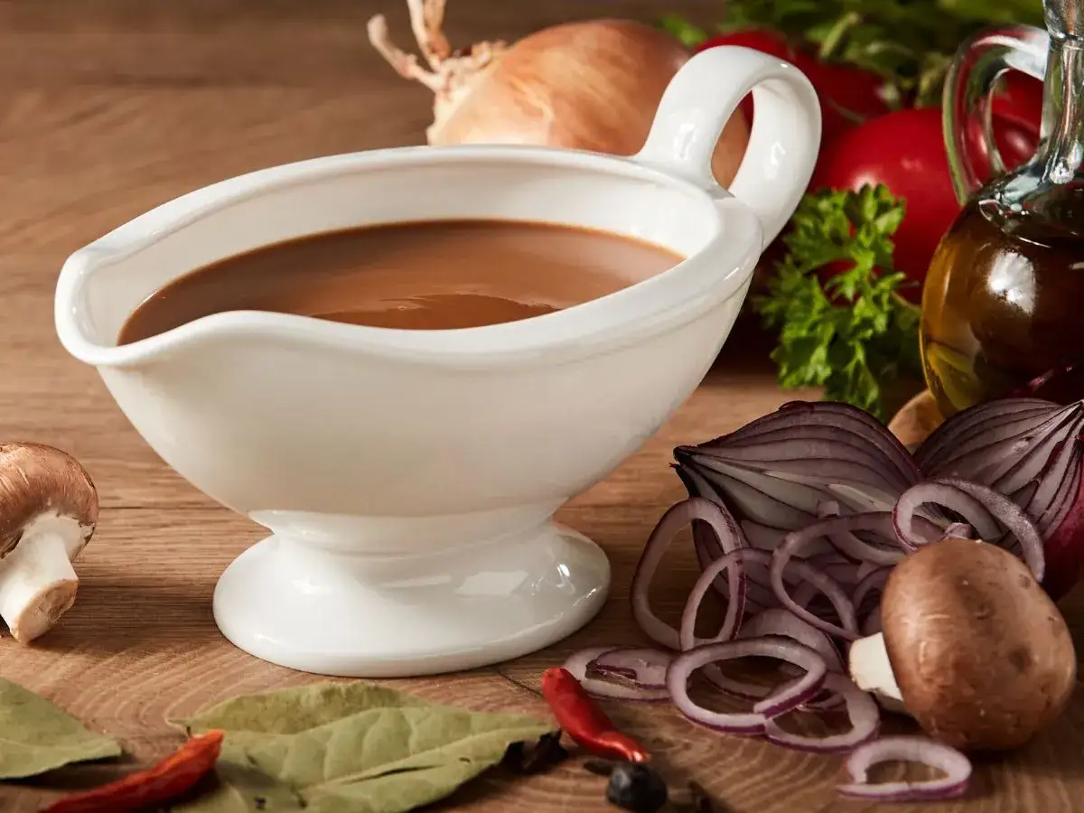 Biała sosjerka z aromatycznym, brązowym sosem. Obok składniki do sosu: cebula, pieczarki, chili. Idealny przepis, jak zrobić pyszny sos.