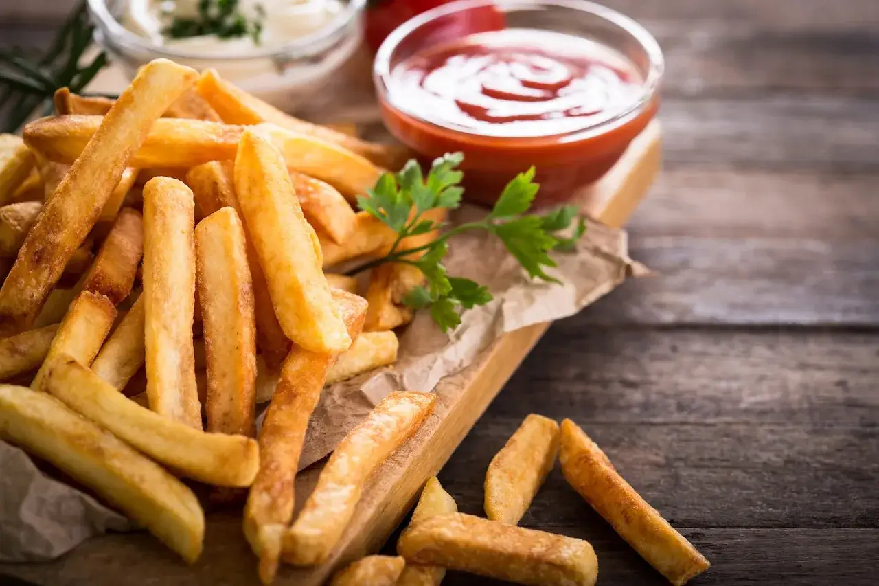 Złociste frytki na desce, obok ketchup i majonez. Ciemnienie frytek podczas smażenia może być spowodowane nadmiarem cukru w ziemniaku.