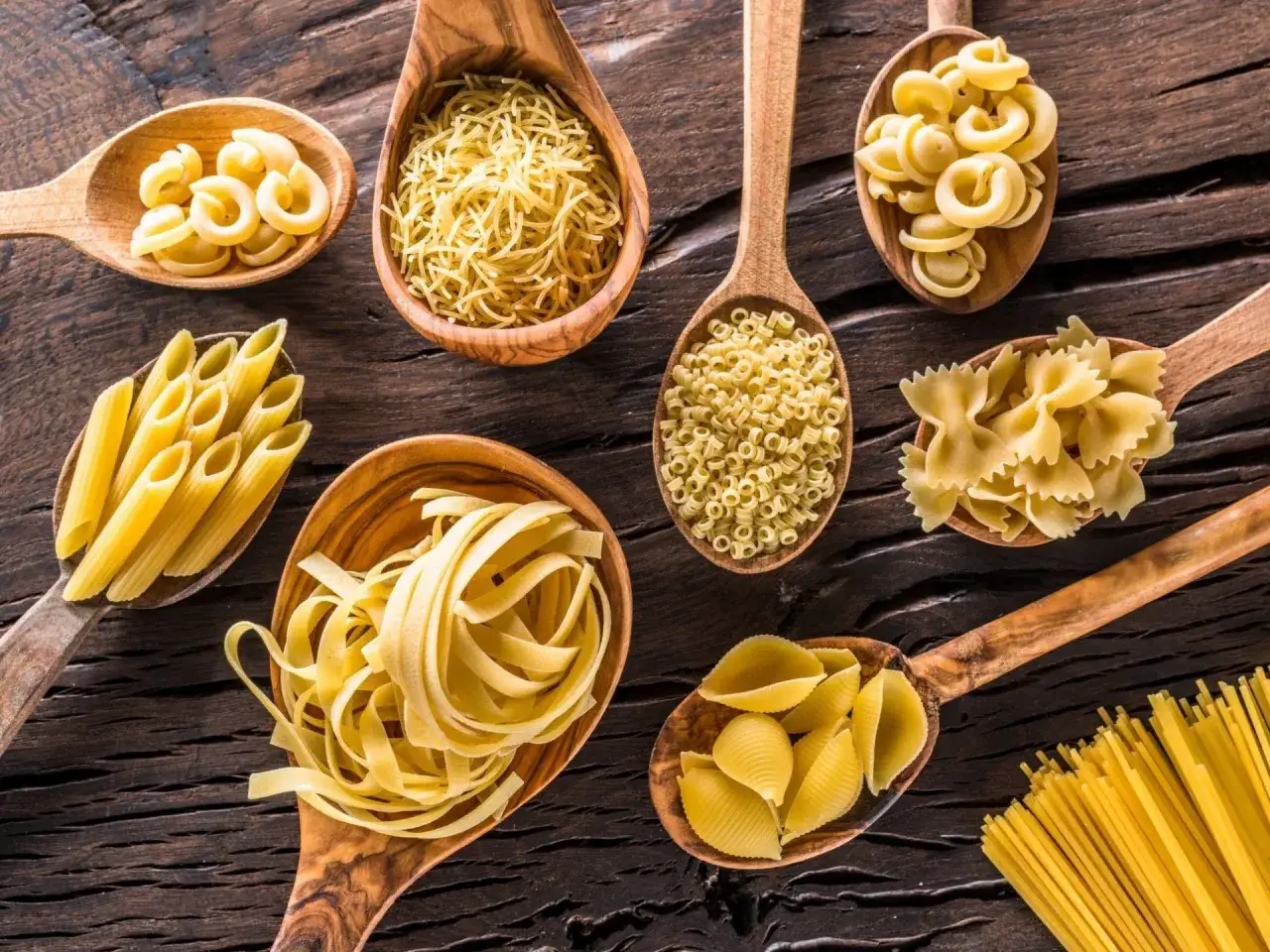 Różne rodzaje makaronów na drewnianych łyżkach: penne, spaghetti, farfalle, conchiglie. Jak dobrać sos do makaronu spaghetti a penne zasady przywierania.
