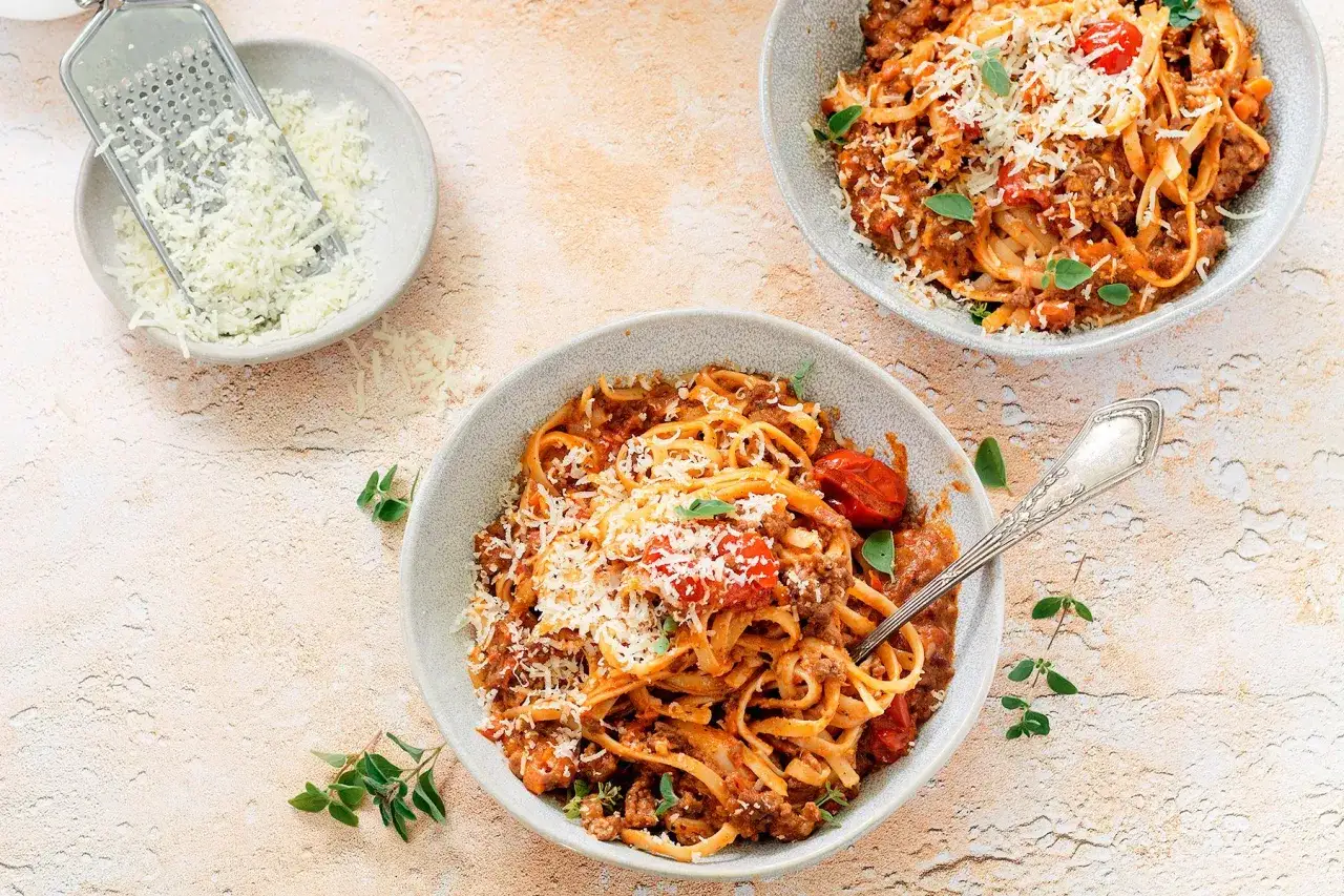 Dwa talerze spaghetti bolognese, posypane parmezanem. Obok miseczka z tartym serem i tarka. Długie gotowanie sosu to klucz do głębi smaku.