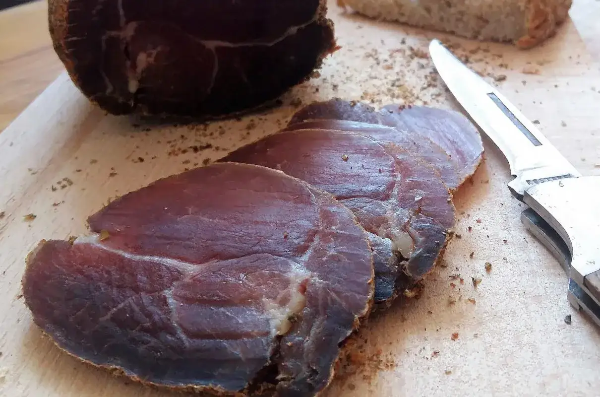 Plastry dojrzewającej szynki, ukazujące różnicę między szynką parmeńską a serrano w procesie dojrzewania. Obok nóż i kromka chleba.