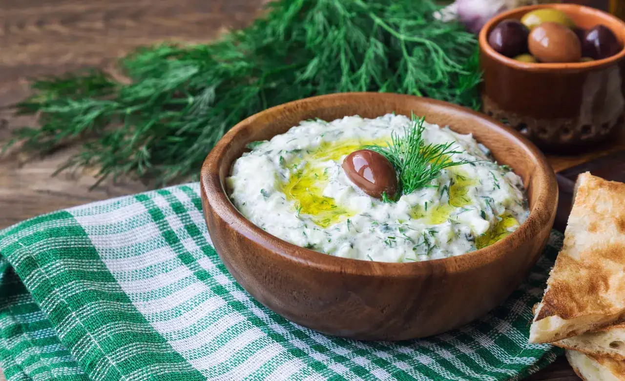 Oryginalny przepis na sos tzatziki: kremowy jogurt, świeży ogórek, czosnek, koperek i oliwa. Podany w drewnianej misce z oliwką i pieczywem.