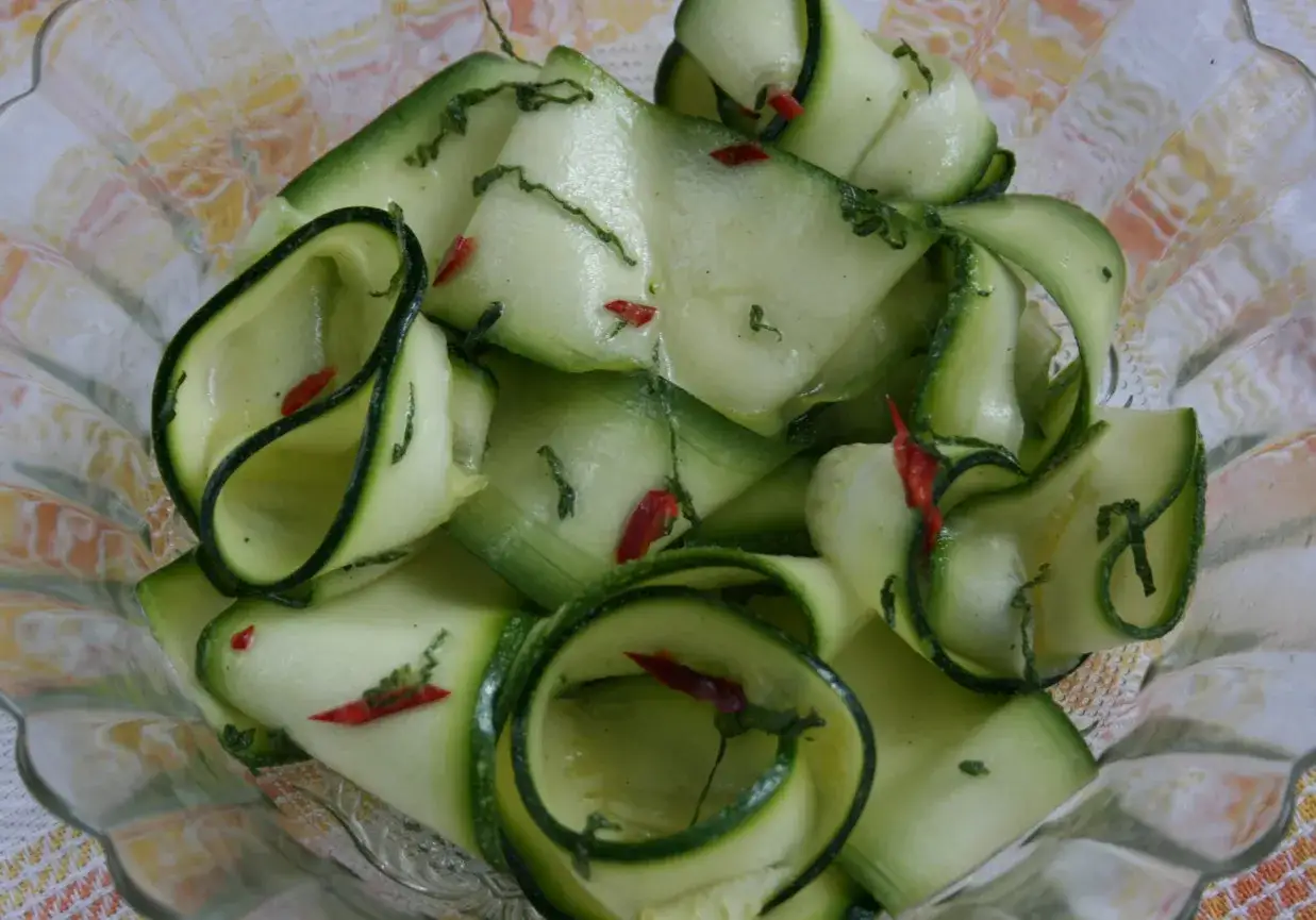 Jak przygotować surówkę z cukinii na surowo w plastrach: cienkie paski cukinii z chili i ziołami w szklanej misce.