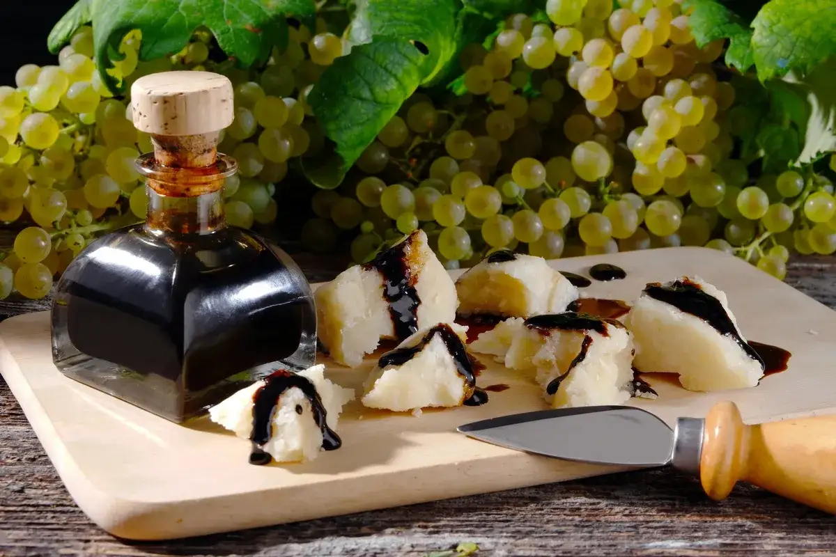 Różnica między octem balsamicznym a kremem balsamicznym w dekoracji: ciemny sos balsamiczny polany na kawałki sera, obok butelki octu i winogron.