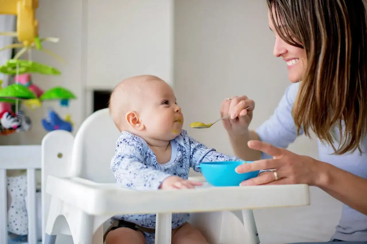 Mama karmi dziecko łyżeczką. W tle widać zabawki. Warto wiedzieć, ile gotować mięso dla niemowlaka, by było zdrowe i smaczne.