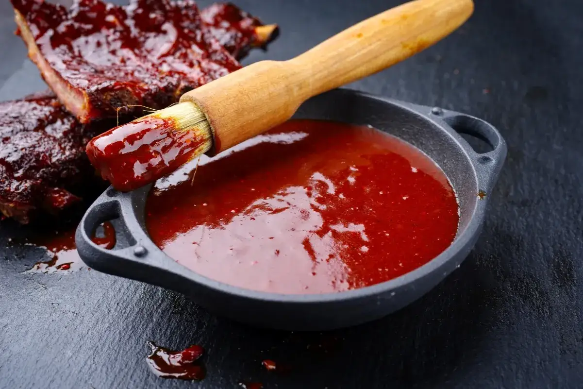 Jak przygotować domowy sos BBQ z wędzonym aromatem bez grilla. Pędzel zanurzony w sosie, obok żeberka w sosie.