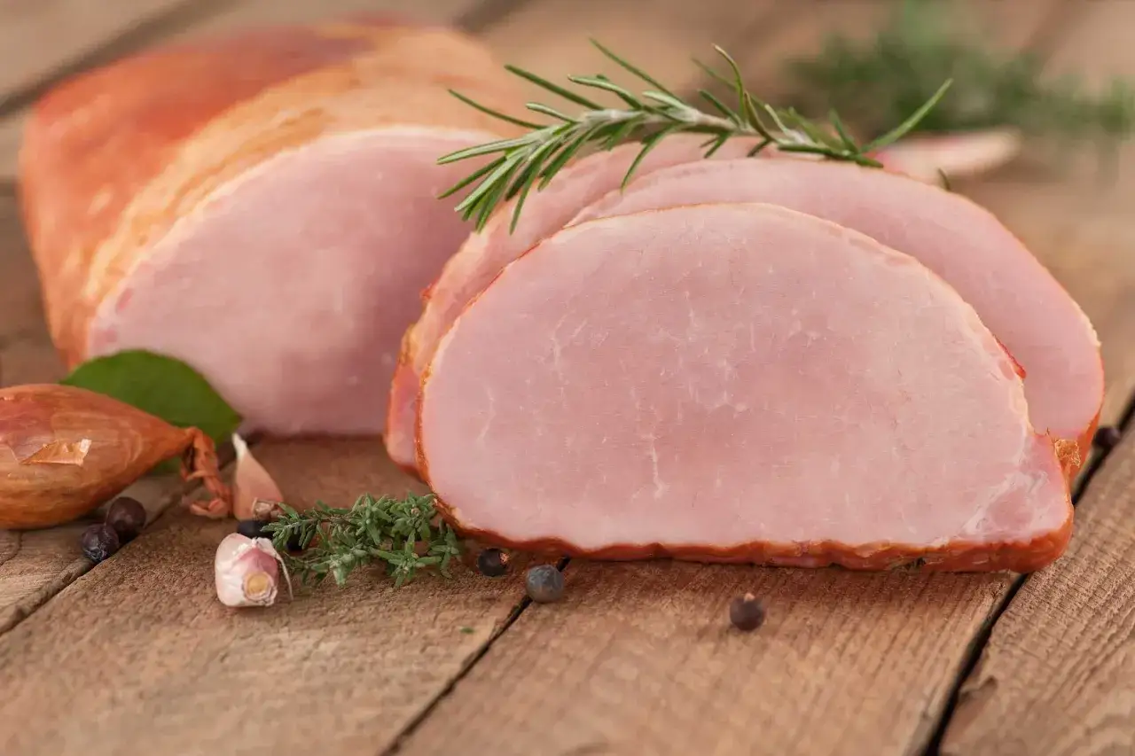 Pyszna, wędzona szynka pokrojona w plastry, z gałązką rozmarynu. Dowiedz się, jak peklować szynkę na mokro, jakie proporcje soli i wody na kilogram wybrać.