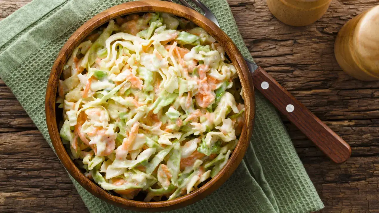 Kremowa sałatka coleslaw z kapusty i marchewki w drewnianej misce. Różnica między sałatką coleslaw a klasyczną surówką z kapusty jest widoczna w kremowym sosie.