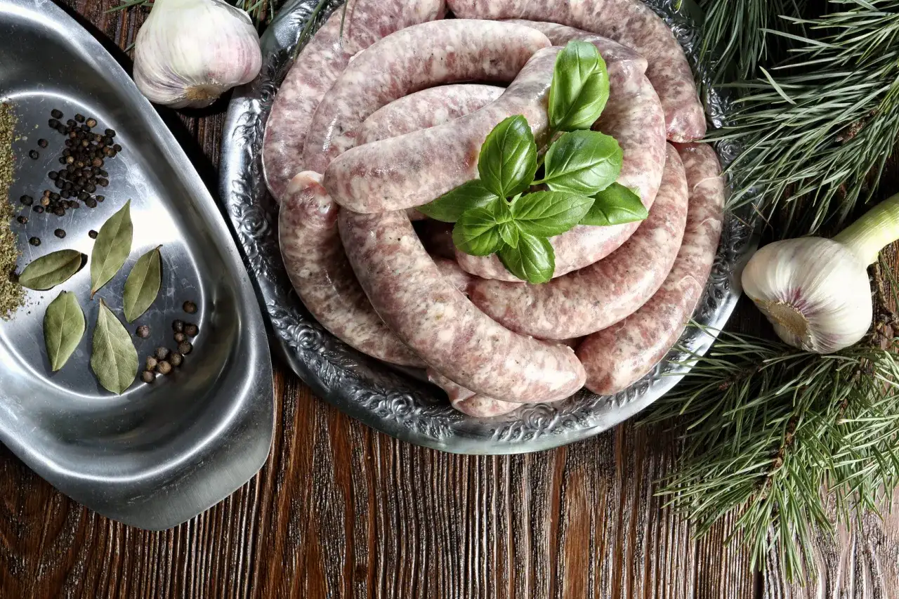 Surowa biała kiełbasa na talerzu, obok czosnek, liście laurowe, pieprz i gałązki sosny. Idealne składniki, by dowiedzieć się, jak sparzyć białą kiełbasę.