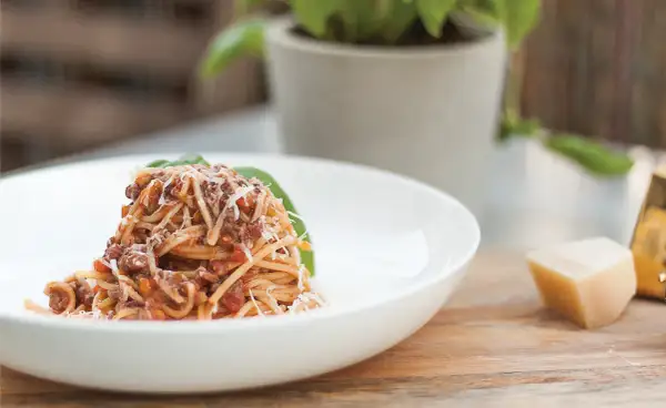 Pyszne spaghetti bolognese przepis oryginalny, podane na białym talerzu, posypane parmezanem. Obok kawałek sera i bazylia.