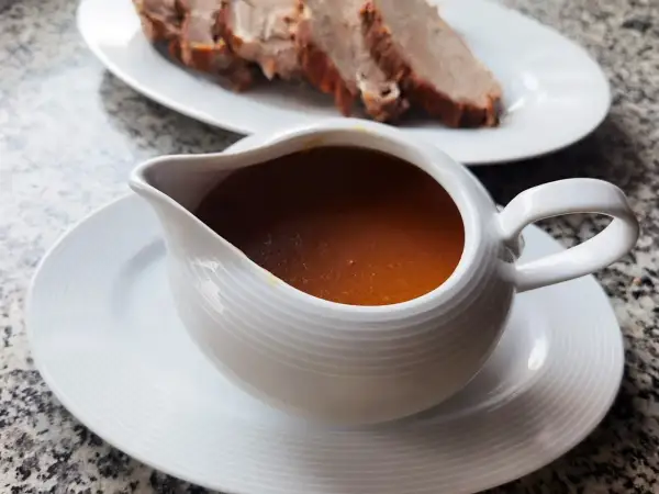 Aromaticzny sos do pieczeni w sosjerce, obok talerz z pokrojonym mięsem. Idealny dodatek, by dowiedzieć się, jak zrobić sos do pieczeni.