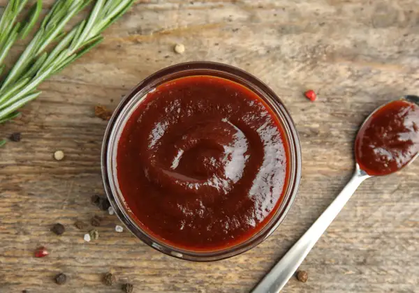 Domowy sos BBQ z wędzonym aromatem bez grilla. Gęsty, ciemny sos w miseczce i na łyżce, obok gałązka rozmarynu i ziarna pieprzu.