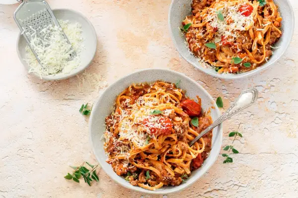 Dwa talerze spaghetti bolognese, posypane parmezanem. Obok miseczka z tartym serem i tarka. Długie gotowanie sosu to klucz do głębi smaku.